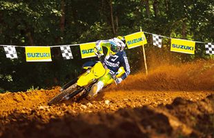 A yellow and blue 2026 Suzuki RM-Z450 dirt bike racer leans sharply into a turn on a dirt track, with a large cloud of orange-brown dust behind the bike and Suzuki banners hanging in the background.