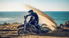A person riding a 2026 Triumph TF 450-E dirt bike on a sandy beach with the ocean in the background, kicking up a large spray of sand.