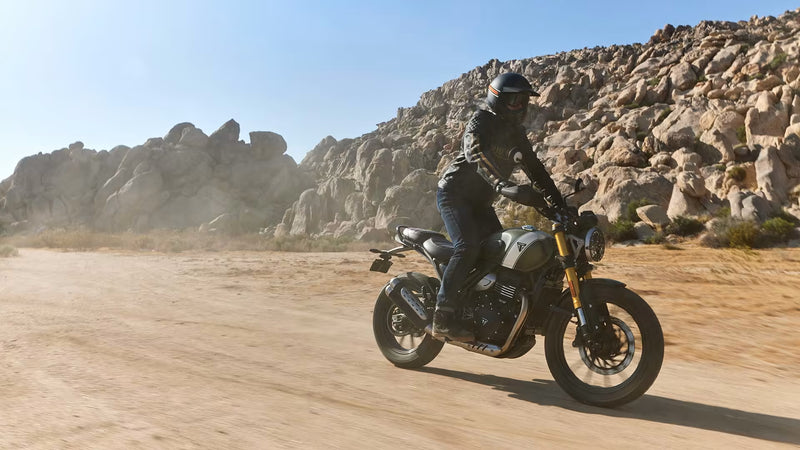 Load image into Gallery viewer, A person wearing a black riding suit and helmet rides a black and gold Triumph Scrambler motorcycle motorcycle on a dusty desert road with large rock formations in the background.
