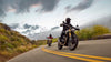 A motorcycle rider in black gear rides a black  Triumph Scrambler 400 motorcycle on a winding road, with another rider and motorcycle visible in the background under a dramatic cloudy sky.